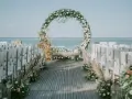 Oto inspiracja, jak zrobić łuk ślubny: okrągła konstrukcja z kwiatów i zieleni nad ławkami przy plaży.