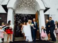 Para młoda wychodzi z kościoła, obsypana konfetti. W tle widać gości, a wśród nich kobietę z prezentem, która może być częścią wykupiny panny młodej.