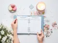 Ręce trzymają otwarty planner ślubny z listą gości i budżetem. Wokół świeca, makaroniki i kwiaty.