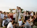 Para Młoda całuje się na tle drewnianego łuku, otoczona gośćmi. Wesele bez dzieci, wszyscy bawią się świetnie.