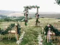 Kwiaty i girlandy zdobią krzesła i bramę weselną, tworząc romantyczną scenerię na ślub w plenerze.