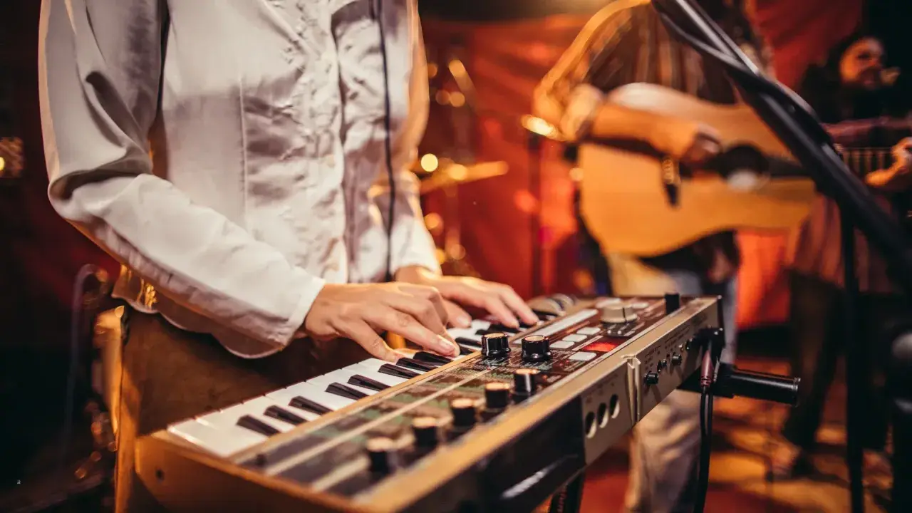 Muzyk gra na keyboardzie, tworząc tło dla najpiękniejszych piosenek biesiadnych. W tle widać gitarzystę.