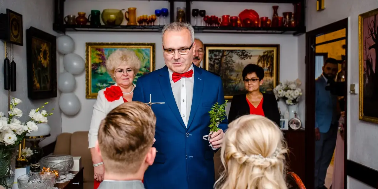 Uroczysty błogosławieństwo przed ślubem cywilnym. Mężczyzna w garniturze trzyma krzyżyk i gałązkę.