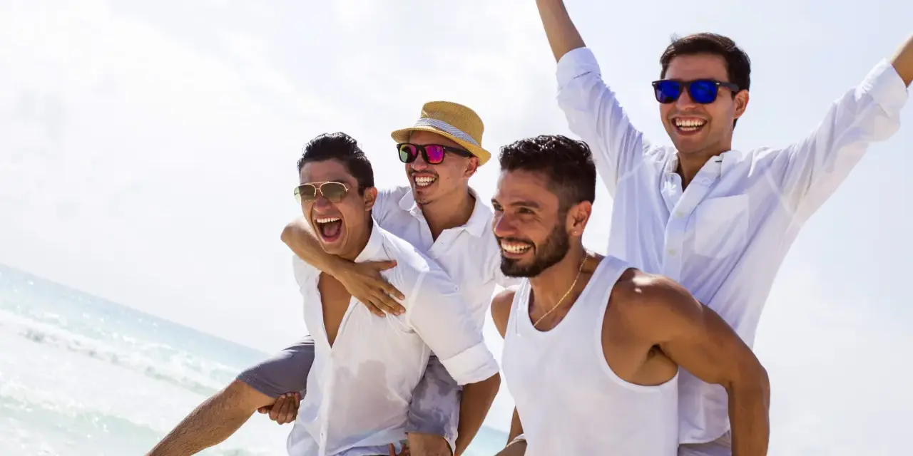 Czterech przyjaciół na plaży, jeden niesie drugiego na barana. Wszyscy uśmiechnięci, w białych koszulach. Wygląda na to, że to oni organizują kawalerski!