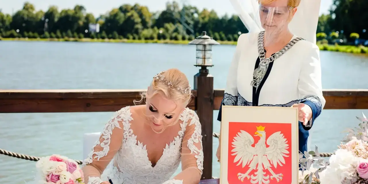 Panna młoda w sukni ślubnej podpisuje dokumenty, obok urzędniczka z herbem Polski. Jej stan cywilny rozwodnik nie przeszkodził w tym radosnym dniu.