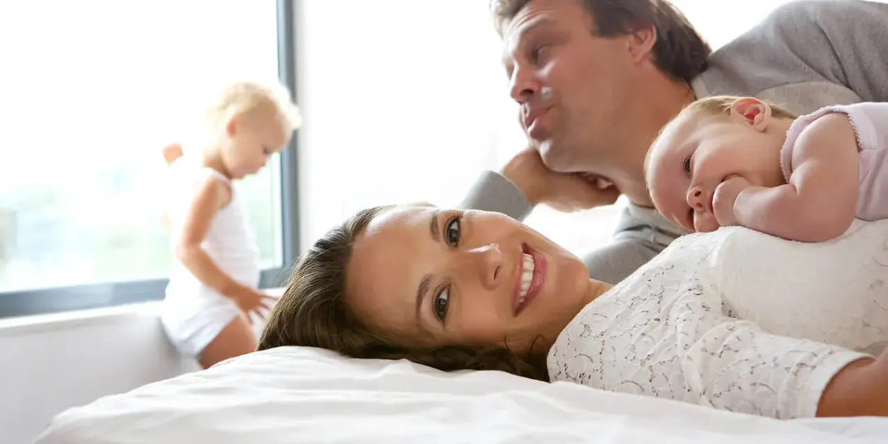 Szczęśliwa rodzina na łóżku. Mama uśmiecha się, dziecko leży na niej, a tata pochyla się nad nimi. W tle bawi się małe dziecko. To idealny obraz na 6 rocznicę ślubu.