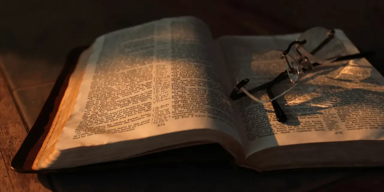 Otwarta Biblia z okularami, oświetlona ciepłym światłem. Co znaczy vel przed nazwiskiem? To skrót od "vel" (łac. "albo"), używany w dokumentach, by wskazać alternatywne nazwisko.