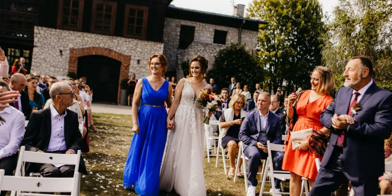 Panna młoda w białej sukni idzie z kobietą w niebieskiej sukience. W tle goście weselni i budynek.