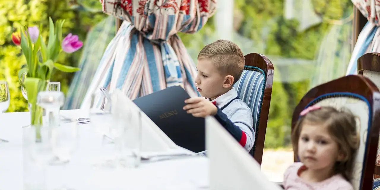 Chłopiec przy weselnym stole dla dzieci przegląda menu. Obok siedzi dziewczynka.