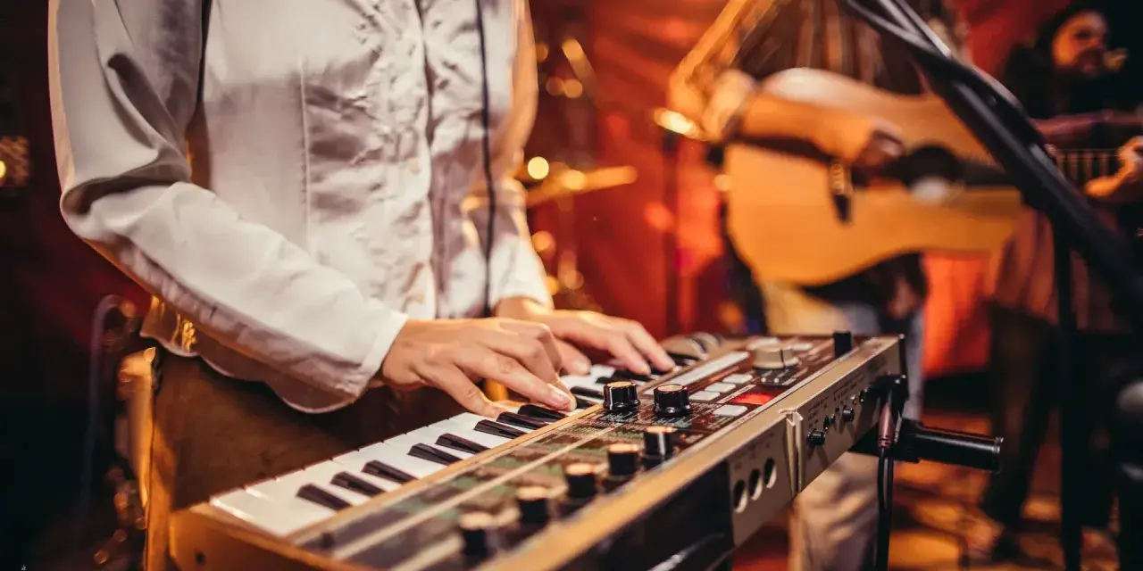 Muzyk gra na keyboardzie, tworząc tło dla najpiękniejszych piosenek biesiadnych. W tle widać gitarzystę.