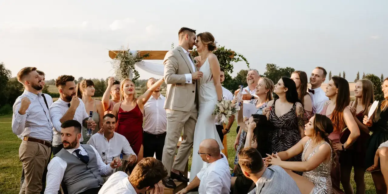Para Młoda całuje się na tle drewnianego łuku, otoczona gośćmi. Wesele bez dzieci, wszyscy bawią się świetnie.