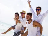 Czterech przyjaciół na plaży, jeden niesie drugiego na barana. Wszyscy uśmiechnięci, w białych koszulach. Wygląda na to, że to oni organizują kawalerski!