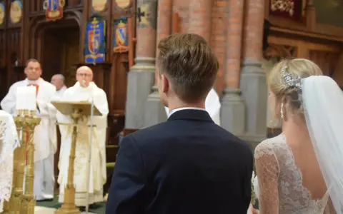 W kościele podczas ślubu, ksiądz czyta wiersze o miłości. Panna młoda w welonie i pan młody w garniturze słuchają.