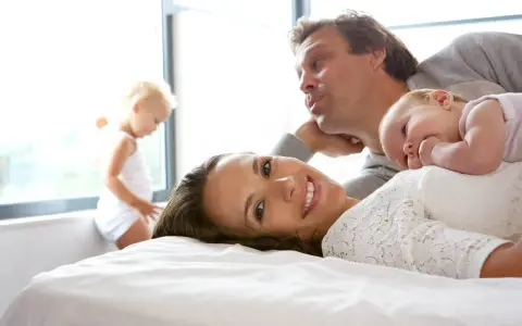 Szczęśliwa rodzina na łóżku. Mama uśmiecha się, dziecko leży na niej, a tata pochyla się nad nimi. W tle bawi się małe dziecko. To idealny obraz na 6 rocznicę ślubu.