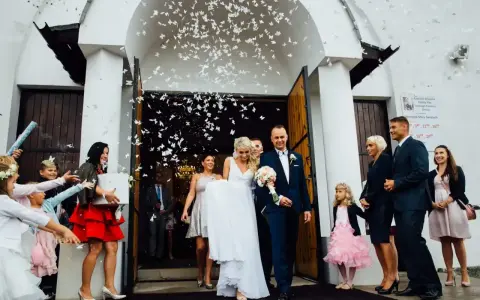 Para młoda wychodzi z kościoła, obsypana konfetti. W tle widać gości, a wśród nich kobietę z prezentem, która może być częścią wykupiny panny młodej.