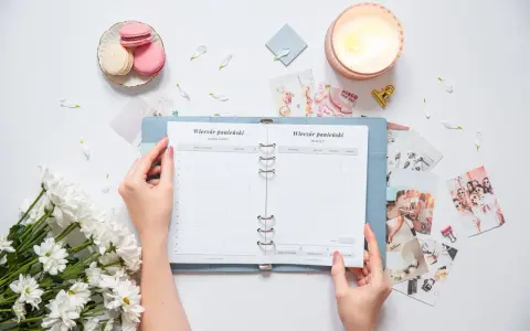 Ręce trzymają otwarty planner ślubny z listą gości i budżetem. Wokół świeca, makaroniki i kwiaty.