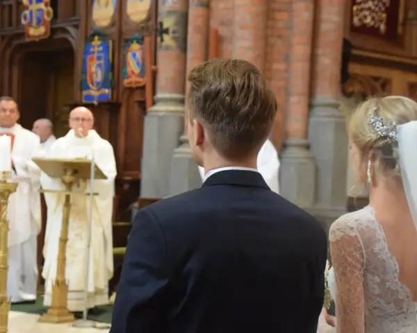 W kościele podczas ślubu, ksiądz czyta wiersze o miłości. Panna młoda w welonie i pan młody w garniturze słuchają.