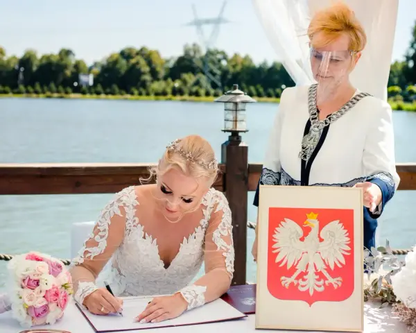 Panna młoda w sukni ślubnej podpisuje dokumenty, obok urzędniczka z herbem Polski. Jej stan cywilny rozwodnik nie przeszkodził w tym radosnym dniu.