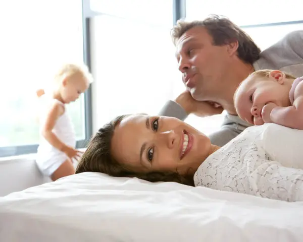 Szczęśliwa rodzina na łóżku. Mama uśmiecha się, dziecko leży na niej, a tata pochyla się nad nimi. W tle bawi się małe dziecko. To idealny obraz na 6 rocznicę ślubu.