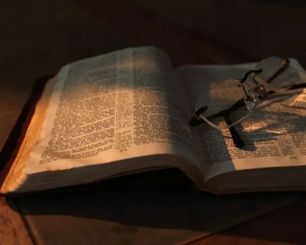 Otwarta Biblia z okularami, oświetlona ciepłym światłem. Co znaczy vel przed nazwiskiem? To skrót od "vel" (łac. "albo"), używany w dokumentach, by wskazać alternatywne nazwisko.