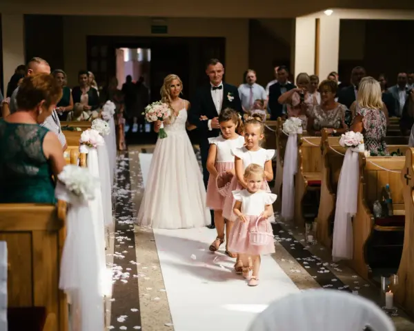 Panna młoda z bukietem idzie do ołtarza w kościele ślubnym, a za nią podążają dziewczynki sypiące kwiaty.