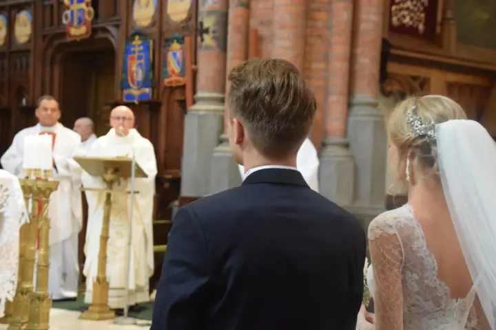 W kościele podczas ślubu, ksiądz czyta wiersze o miłości. Panna młoda w welonie i pan młody w garniturze słuchają.
