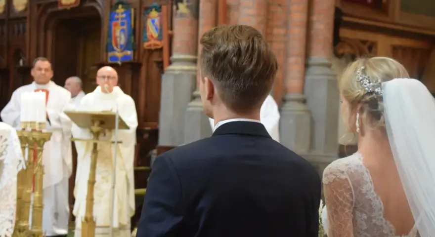 W kościele podczas ślubu, ksiądz czyta wiersze o miłości. Panna młoda w welonie i pan młody w garniturze słuchają.