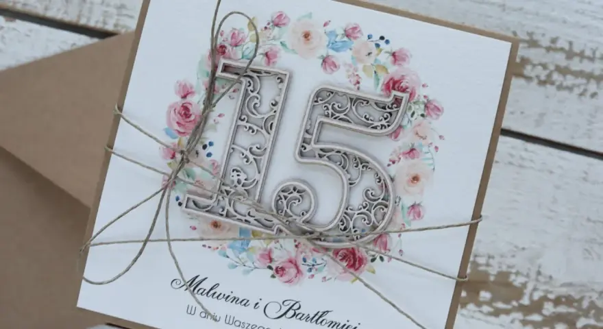 Elegancka kartka z napisem "Malwina i Bartłomiej w dniu Waszego Jubileuszu" i ozdobnym numerem "15" na tle kwiatów. To zaproszenie na 15 rocznicę ślubu, zwaną rocznicą szklaną.
