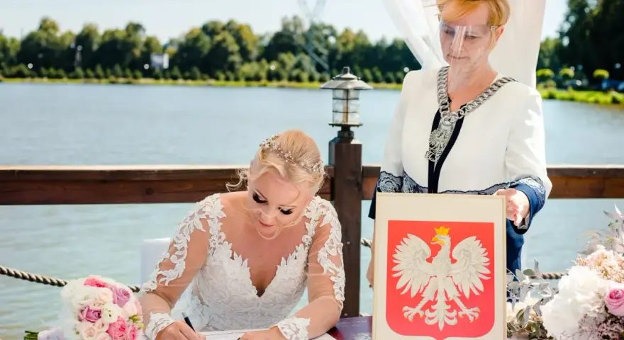 Panna młoda w sukni ślubnej podpisuje dokumenty, obok urzędniczka z herbem Polski. Jej stan cywilny rozwodnik nie przeszkodził w tym radosnym dniu.