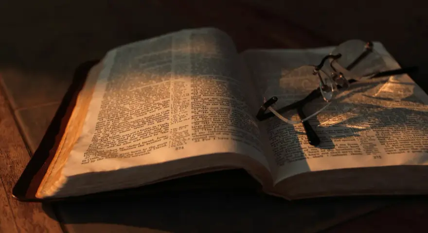 Otwarta Biblia z okularami, oświetlona ciepłym światłem. Co znaczy vel przed nazwiskiem? To skrót od "vel" (łac. "albo"), używany w dokumentach, by wskazać alternatywne nazwisko.