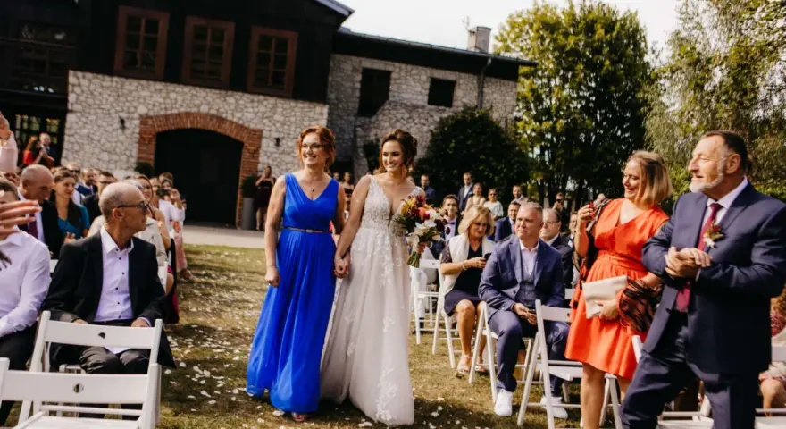 Panna młoda w białej sukni idzie z kobietą w niebieskiej sukience. W tle goście weselni i budynek.