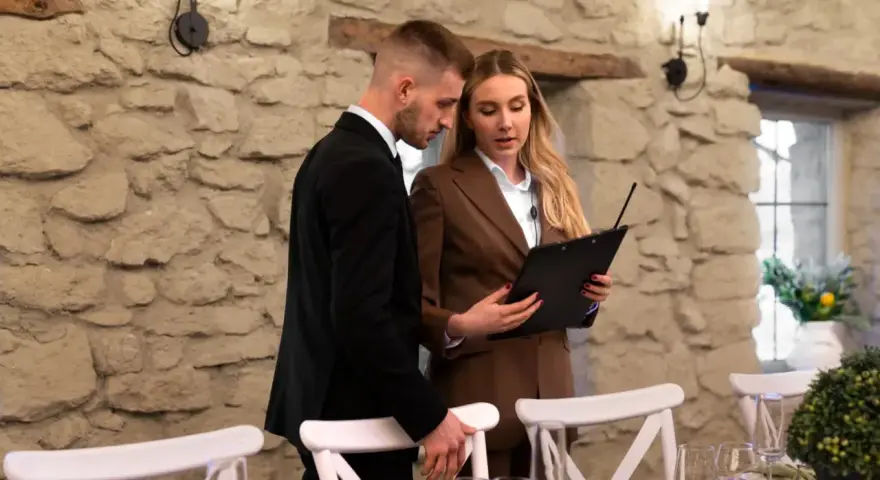 Para weselnych planuje, jak usadzić gości na weselu, przeglądając listę przy kamiennej ścianie.