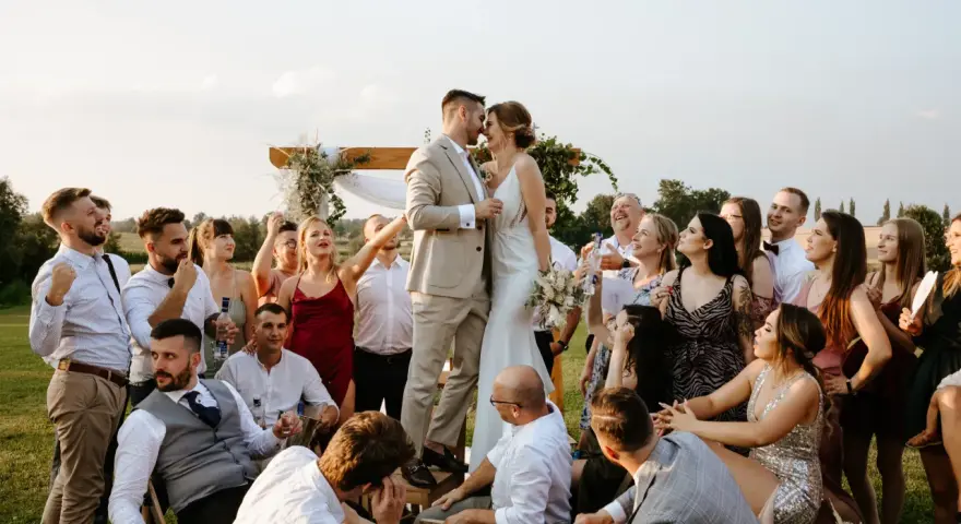Para Młoda całuje się na tle drewnianego łuku, otoczona gośćmi. Wesele bez dzieci, wszyscy bawią się świetnie.