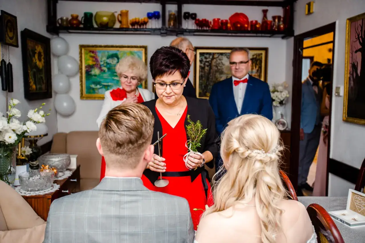 Wierszyk na błogosławieństwo młodej pary od dziecka. Kobieta w czerwonej sukience trzyma gałązkę, błogosławiąc narzeczonych.