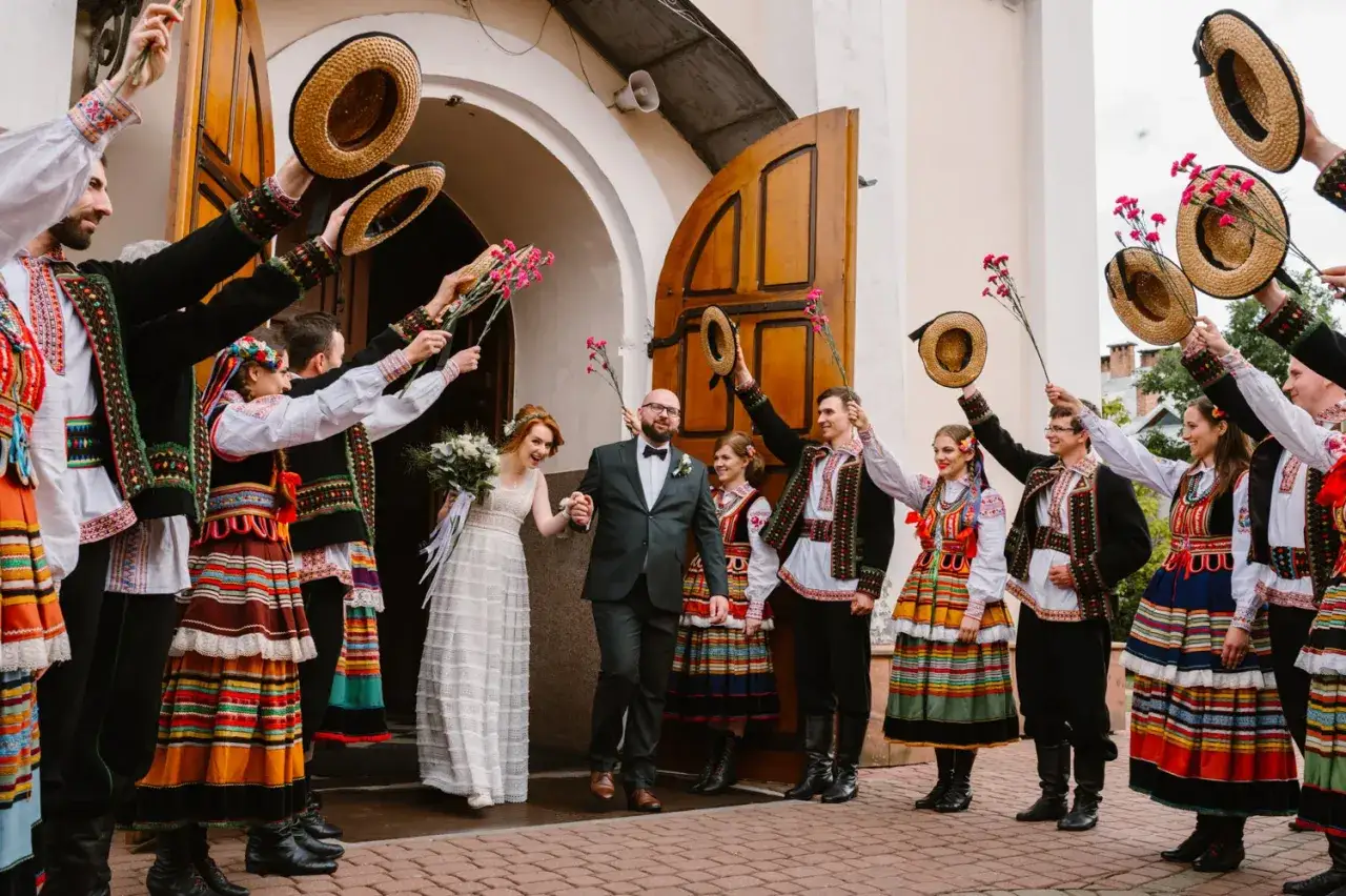 Para młoda wychodzi z kościoła, witana przez gości w strojach ludowych. Wszyscy trzymają w g&oacute;rze słomiane kapelusze i kwiaty, celebrując słowiańskie wesele.