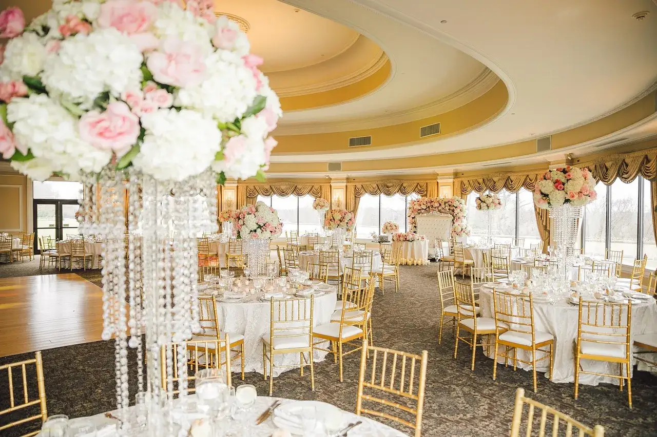Elegancka sala weselna z białymi i różowymi kwiatami, złotymi krzesłami i kryształowymi żyrandolami. Tak wygląda wesele, co to za piękna uroczystość!