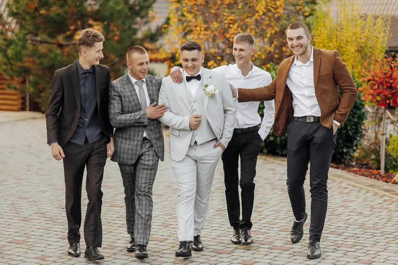 Pięciu mężczyzn w eleganckich garniturach, idealny dress code na wesele. Pan młody w jasnym garniturze, druhowie w ciemniejszych stylizacjach.