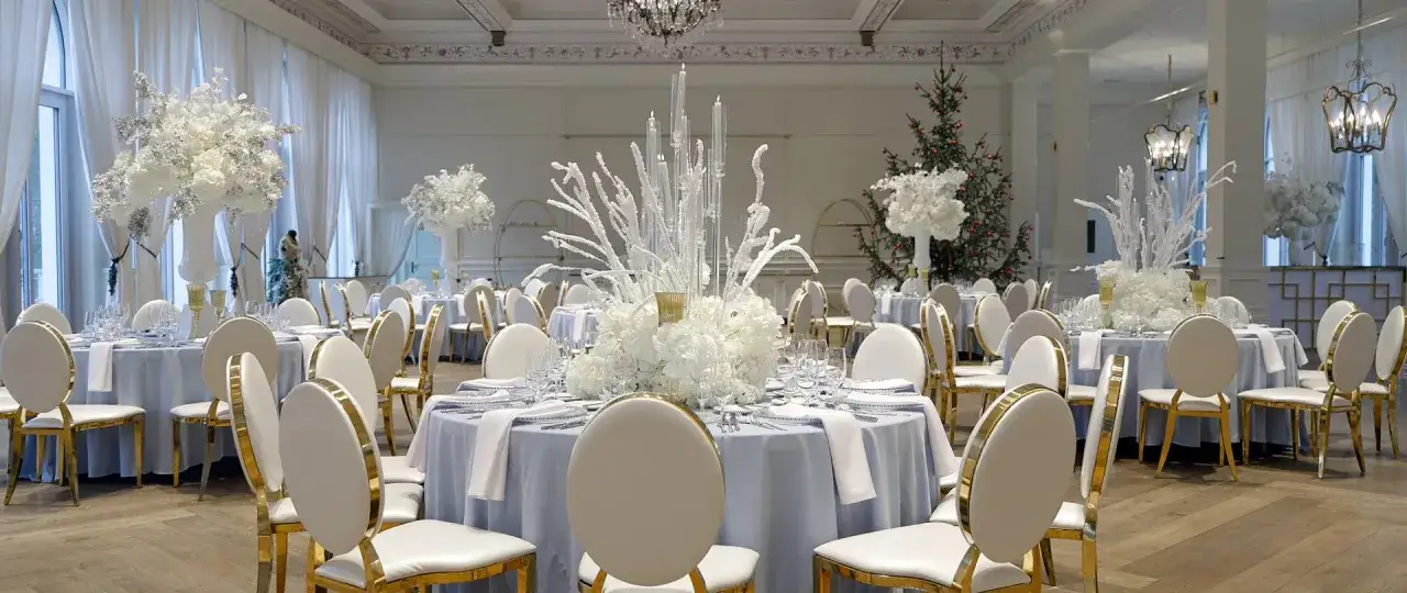 Eleganckie przyjęcie weselne zimą. Białe kwiaty i świece na stołach, złote krzesła i choinka w tle tworzą magiczną atmosferę.